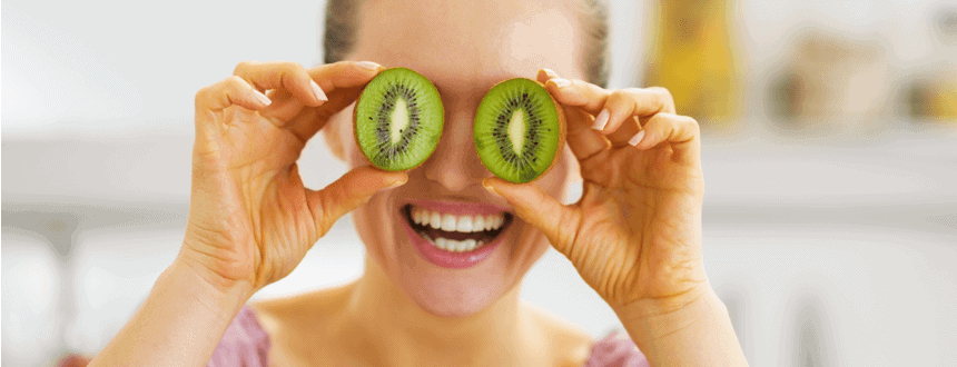 Smiling young woman holding kiwi slices in front of eyes